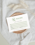 A printed gift voucher card for McQuarrie Skin & Beauty, styled on a round wooden coaster over a textured white fabric backdrop with dried grasses.