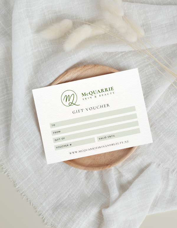 A printed gift voucher card for McQuarrie Skin & Beauty, styled on a round wooden coaster over a textured white fabric backdrop with dried grasses.